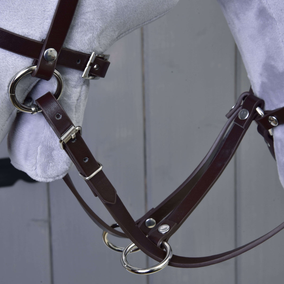 Close-up of the Martingale brown by Hobby Horse LarDen, featuring a brown leather bridle with metal buckles and rings on a light grey model horse head, set against a grey wooden background.