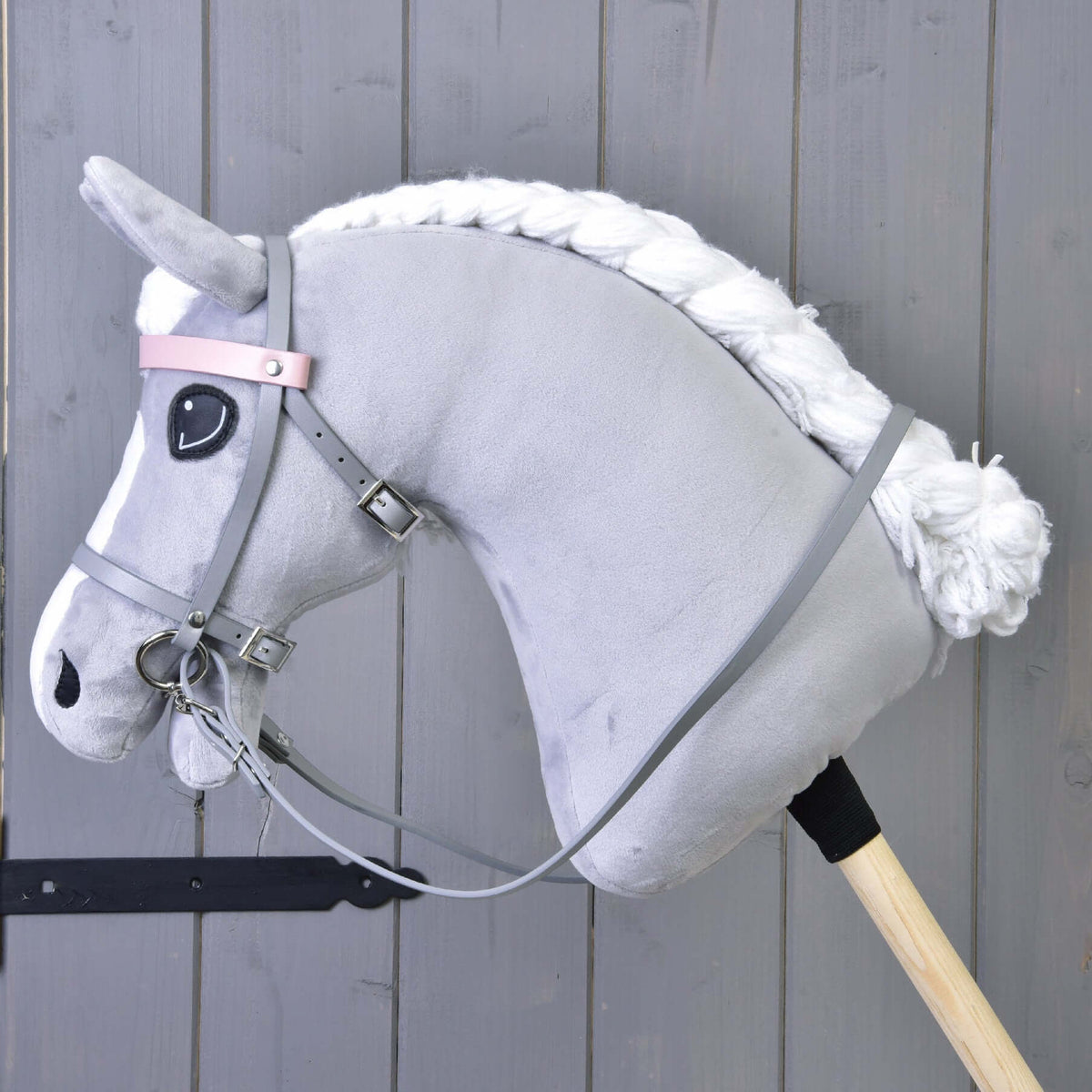 The Hobby Horse LarDen gray plush hobby horse with a white yarn mane, black eyes, and a Grey leatherette set—bridle, reins, and martingale—is mounted on a wooden stick against a gray wooden wall background.