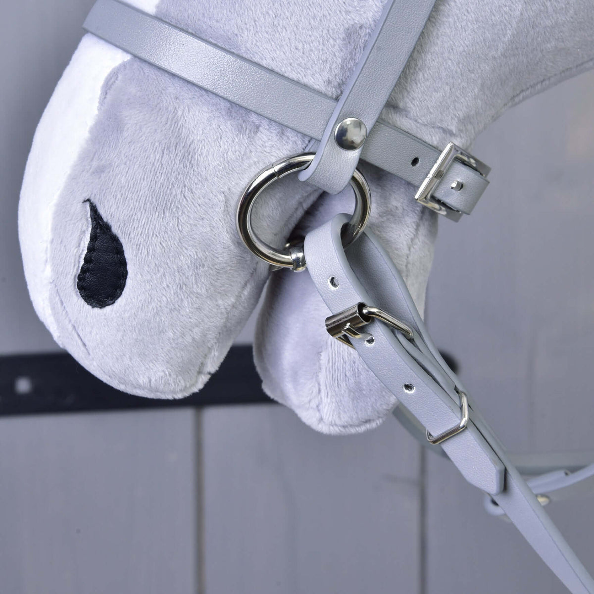 Close-up of Hobby Horse LarDen's gray plush hobby horse wearing the Grey leatherette set—bridle, reins, and martingale with metal rings and buckles. Features stitched black eyes; blurred wood paneling in background.