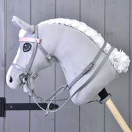 A Hobby Horse LarDen plush gray horse with a white mane features a Grey leatherette set—bridle, reins, and martingale—mounted on a wooden stick in front of a gray wooden wall.