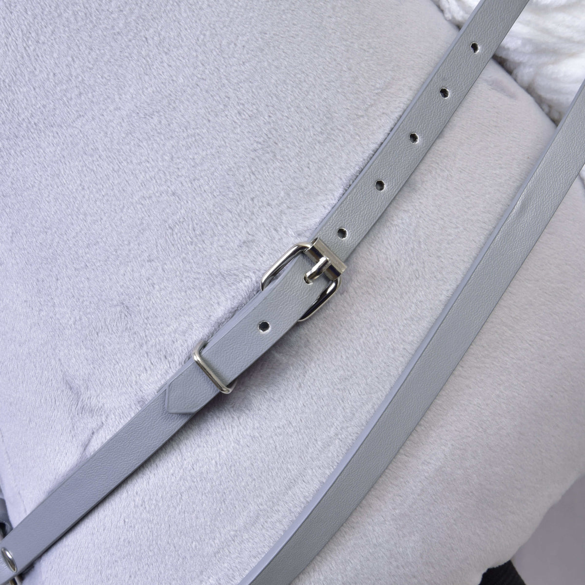 A close-up of the Grey leatherette set—bridle, reins, and martingale by Hobby Horse LarDen—showing the light gray strap with a metal buckle on a pale gray fabric, ideal for enhancing your hobby horse tack.