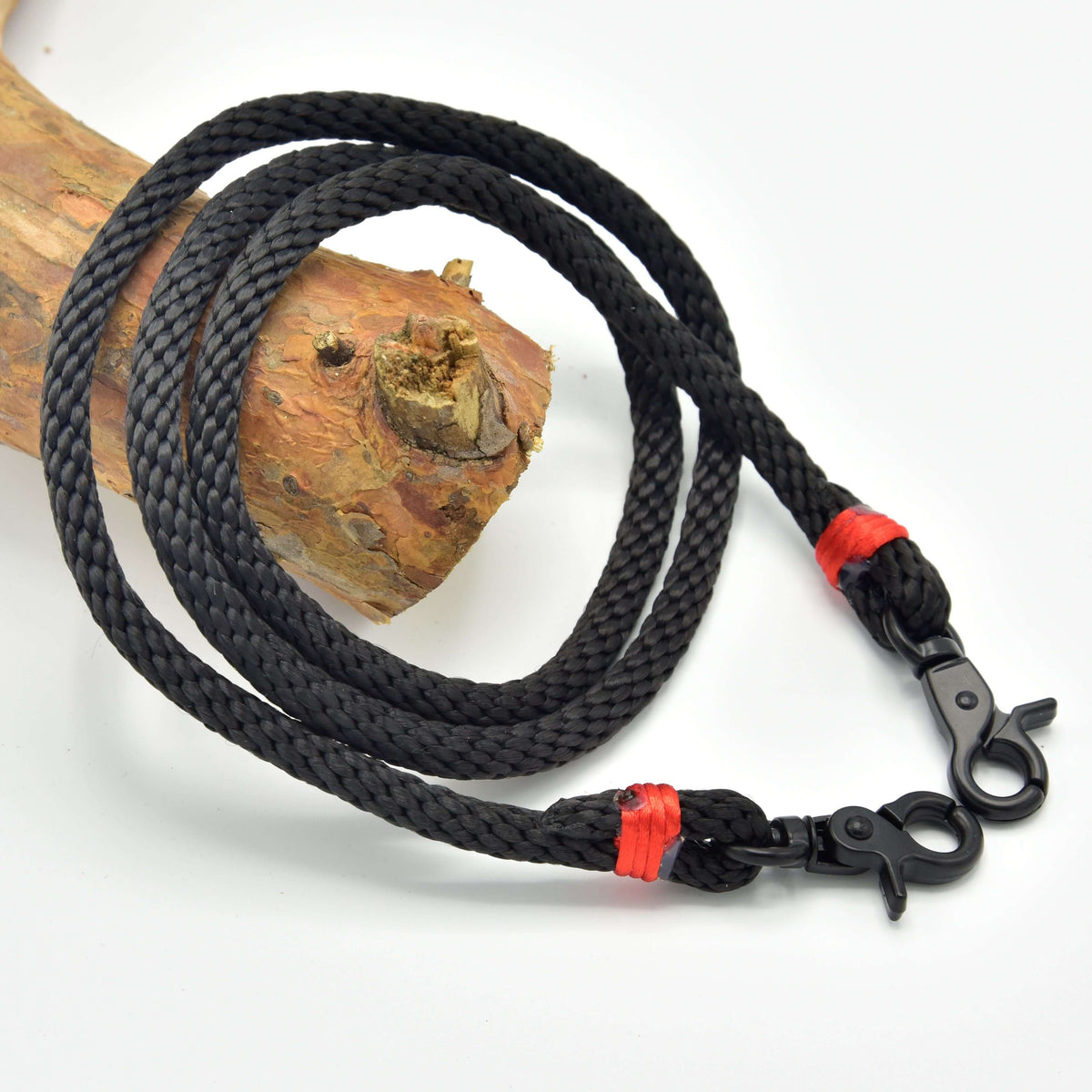 The Reins Spark by Hobby Horse LarDen features black braided polypropylene rope with metal clips at both ends and red binding, coiled on a wooden branch—ideal as handmade reins or hobby horse reins.