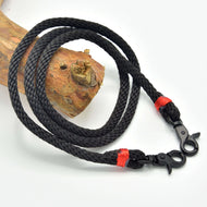 The Reins Spark by Hobby Horse LarDen features black braided polypropylene rope with metal clips at both ends and red binding, coiled on a wooden branch—ideal as handmade reins or hobby horse reins.